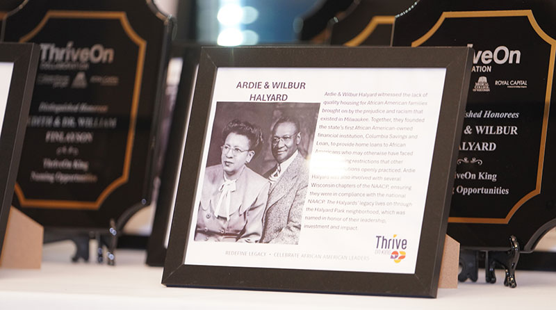 A framed display honoring Ardie and Wilbur Halyard with a black-and-white photo and text about their contributions, with other plaques in the background.